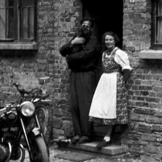 Vintage photo of couple standing by brick house with motorcycle and bicycle.