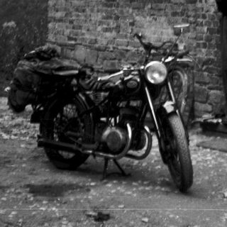 Vintage photo of couple standing by brick house with motorcycle and bicycle.