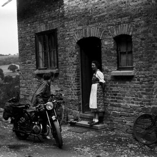 Vintage motorcycle with rider and woman standing by brick house.