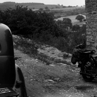 Vintage motorcycle with rider and woman standing by brick house.