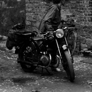 Vintage motorcycle with rider and woman standing by brick house.
