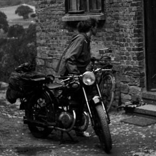 Vintage motorcycle with rider and woman standing by brick house.