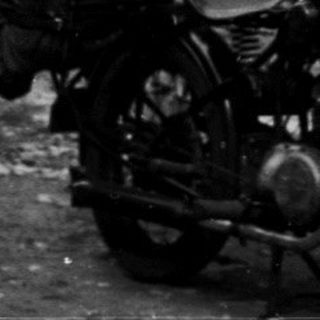 Vintage motorcycle with rider and woman standing by brick house.