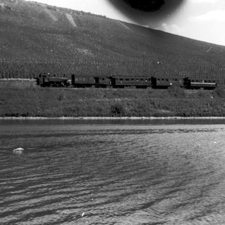 Steam train crossing snowy, mountainous landscape near water.