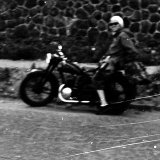 Person riding vintage motorcycle on paved road near stone wall.