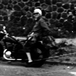 Person riding vintage motorcycle on paved road near stone wall.