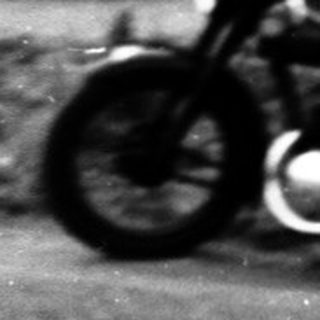 Person riding vintage motorcycle on paved road near stone wall.