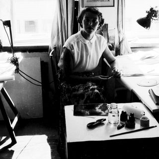 Woman seated at sewing table with fabric and tools.