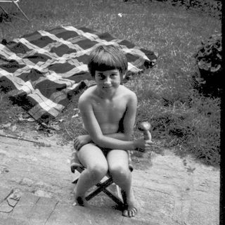Shirtless boy sitting outside on left, backyard scene with umbrella and chairs.