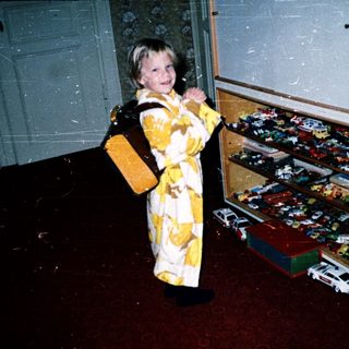 Young child in yellow outfit near toy-filled cabinet.