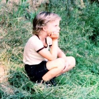 A child sitting alone by a stream in a wooded area.
