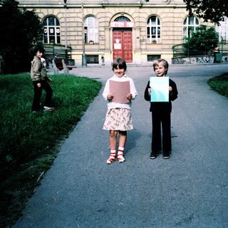 Two children holding large blank papers outdoors near a building.