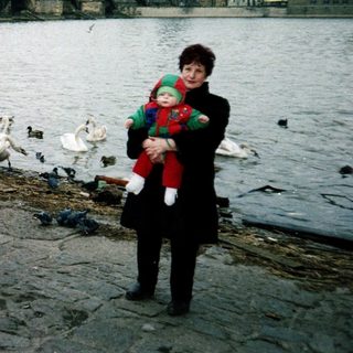 Two women by river with ducks, holding child, Prague bridge in background.