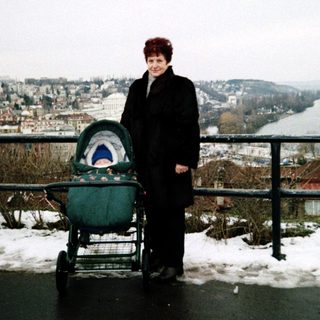 Woman in winter attire with stroller by snowy riverside view.