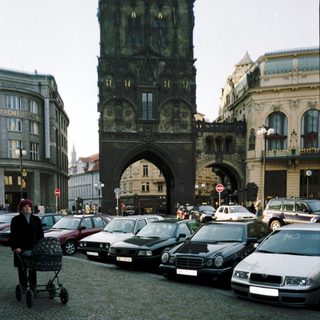 Historic city gate with parked cars and pedestrian pushing stroller.