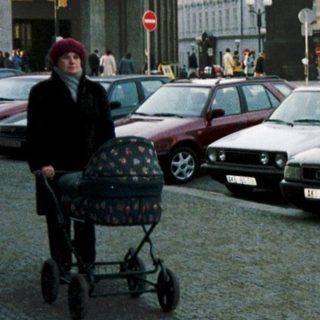 Historic city gate with parked cars and pedestrian pushing stroller.