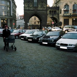 Historic city gate with parked cars and pedestrian pushing stroller.
