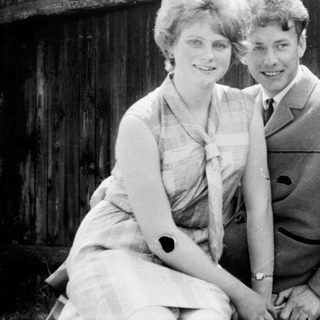 Two people posing happily outdoors near wooden fence.