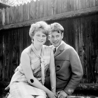 Two people posing happily outdoors near wooden fence.