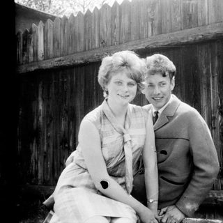 Two people posing happily outdoors near wooden fence.