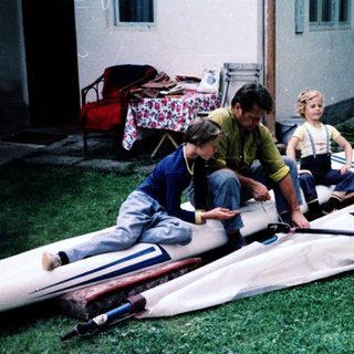 Three children assembling a windsurfing board outdoors.