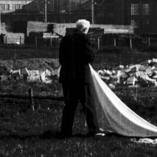 Man holding large white sheet near industrial building.