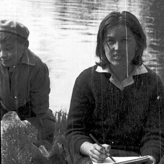 Woman writing by lake with companion in background.