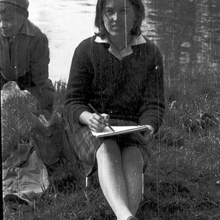Woman writing by lake with companion in background.