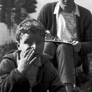 Two men sitting outdoors near water, one reading.