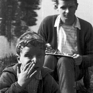 Two men sitting outdoors near water, one reading.