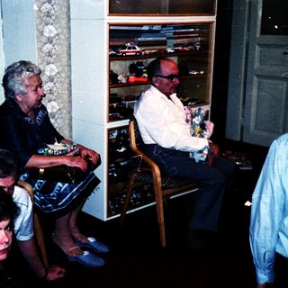 Group of people gathered indoors, seated and standing, watching a display of vintage cars.
