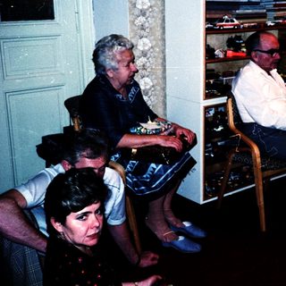 Group of people gathered indoors, seated and standing, watching a display of vintage cars.
