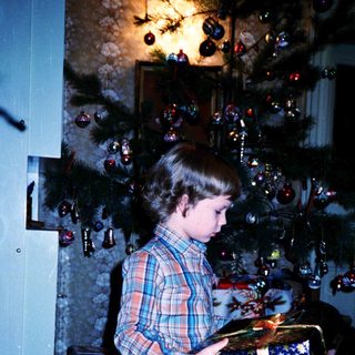 Child opening wrapped gift under decorated Christmas tree.