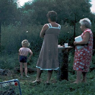 Family gathering around outdoor campfire in grassy field.