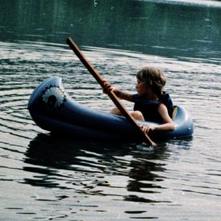 Children paddling inflatable boats on calm lake.