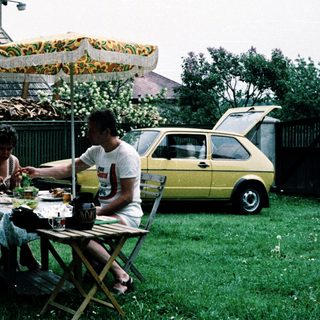 Two people eating outdoors under umbrella near vintage car.