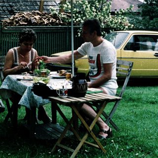Two people eating outdoors under umbrella near vintage car.