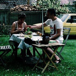 Two people eating outdoors under umbrella near vintage car.