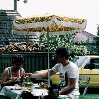 Two people eating outdoors under umbrella near vintage car.