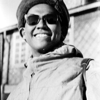 Young person smiling outdoors wearing sunglasses, beret, and backpack.