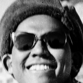 Young person smiling outdoors wearing sunglasses, beret, and backpack.