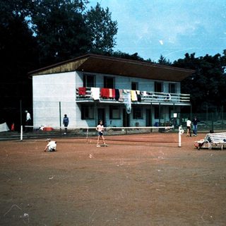 Outdoor tennis court with players and nearby building.