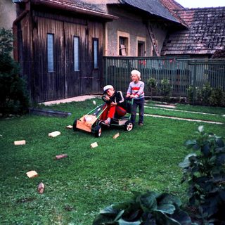 Two people mowing grass with a child in a cart.