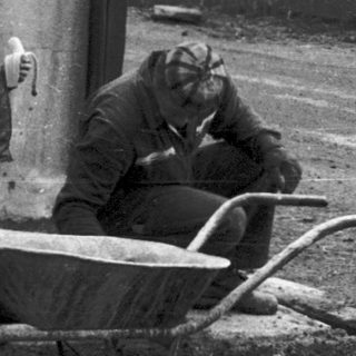 Two men pushing wheelbarrow near concrete structure.
