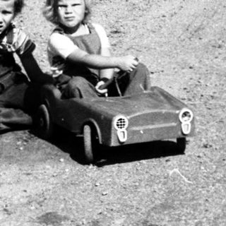 Two children playing with toy cars on gravel road.
