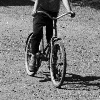 A boy rides a bicycle in a gravel driveway near vintage cars.