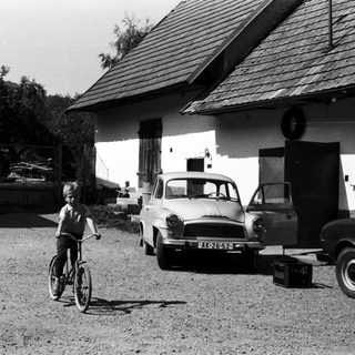 A boy rides a bicycle in a gravel driveway near vintage cars.