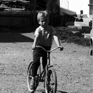 A boy rides a bicycle in a gravel driveway near vintage cars.