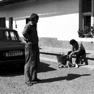 Two men outdoors, one bending to pet dog.