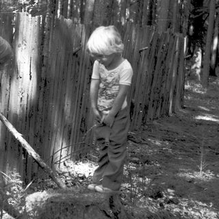 Two children chopping wood outdoors near fence.